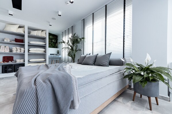 A modern bedroom with a large bed, a built-in wardrobe, a bedside plant, and a view of the city through large windows. - 31 Doors Hotel (Alexandroupoli)