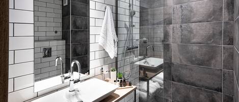 Une salle de bain moderne avec un grand miroir, un lavabo blanc, un meuble-lavabo en bois, une douche avec une porte en verre et des carreaux gris foncé.
