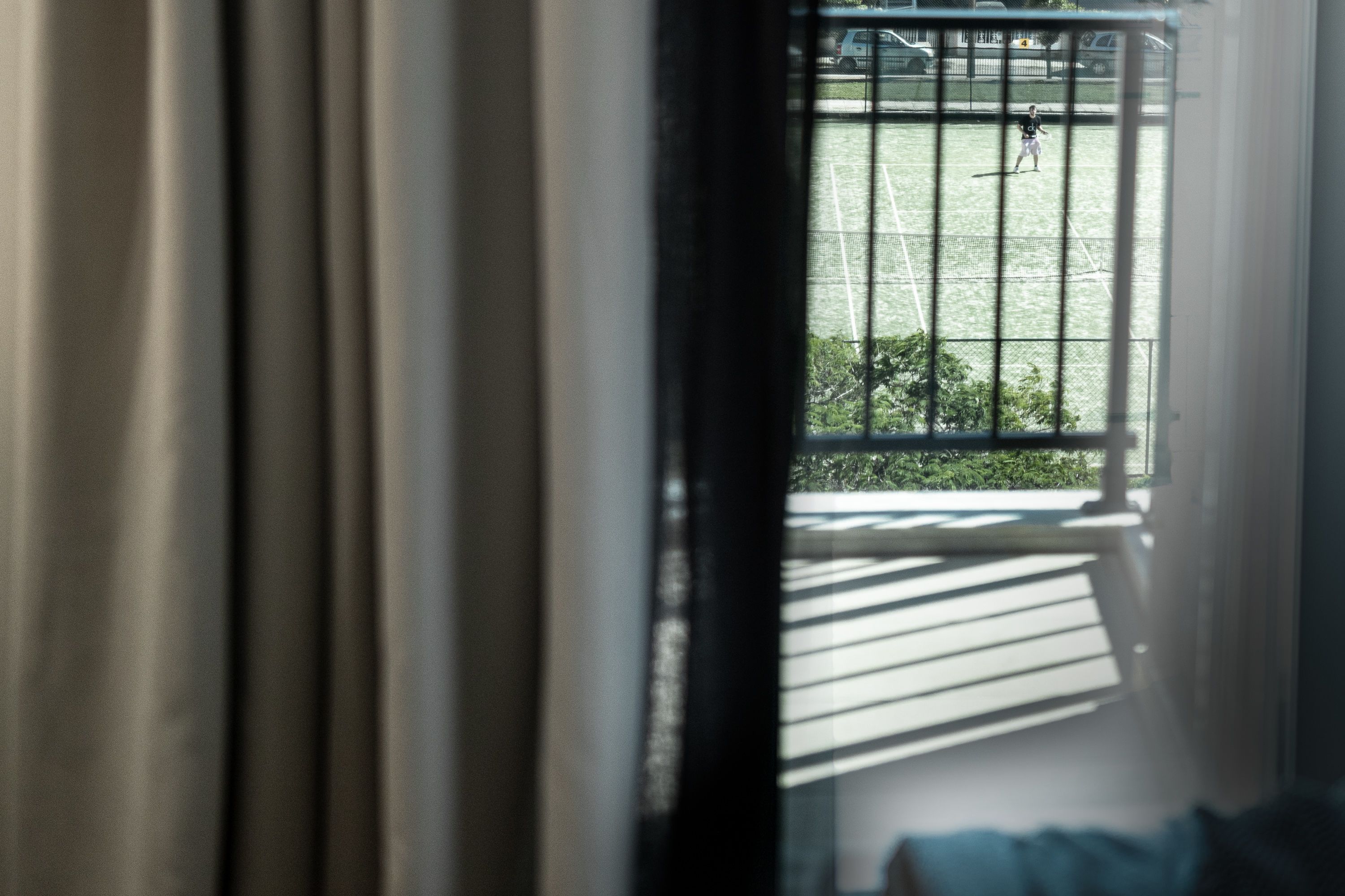 A view through a window with metal bars, a tennis court, and greenery outside.