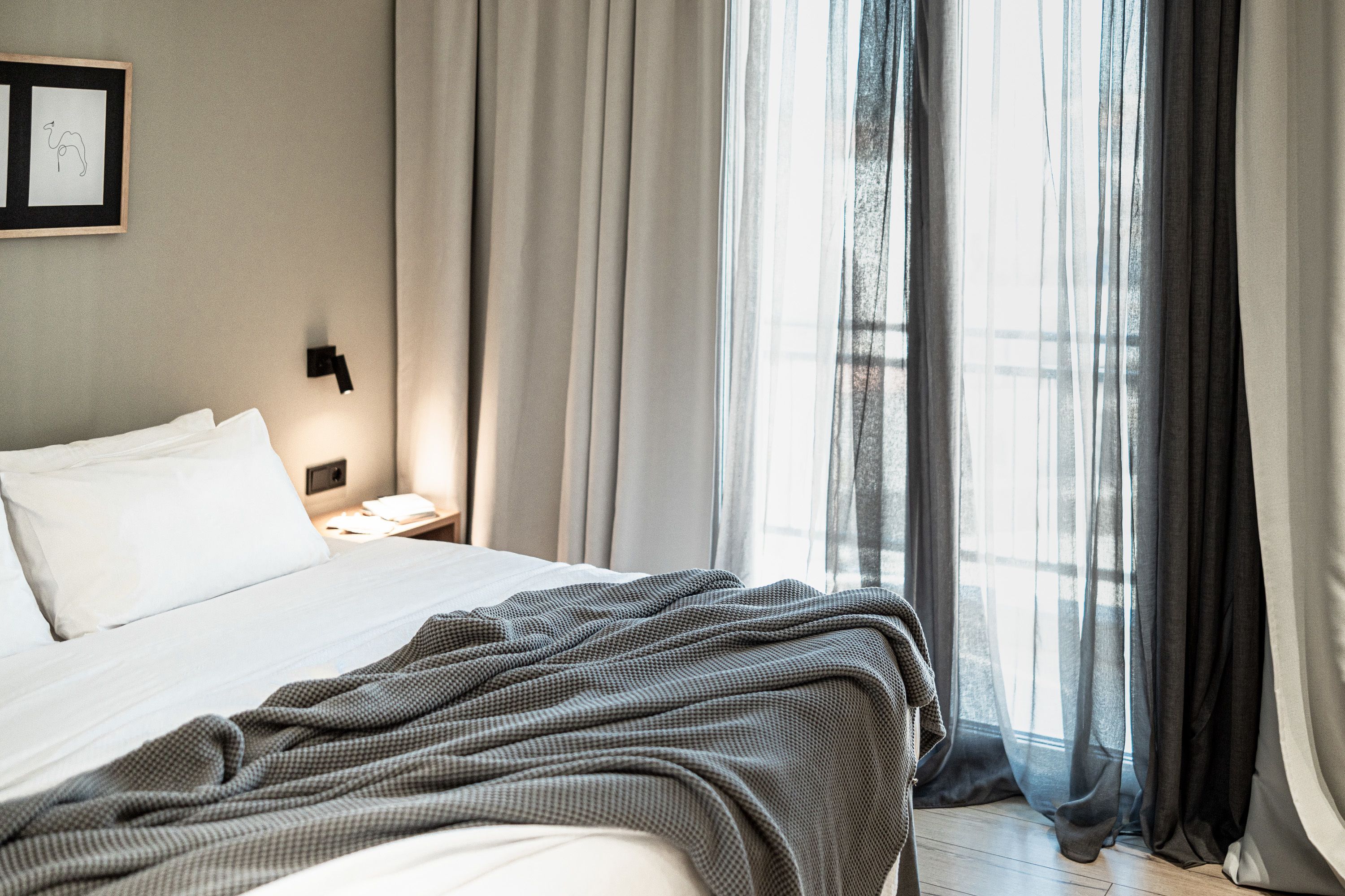 A neatly made bed with a checkered blanket, a bedside table with a lamp, and sheer curtains allowing natural light.