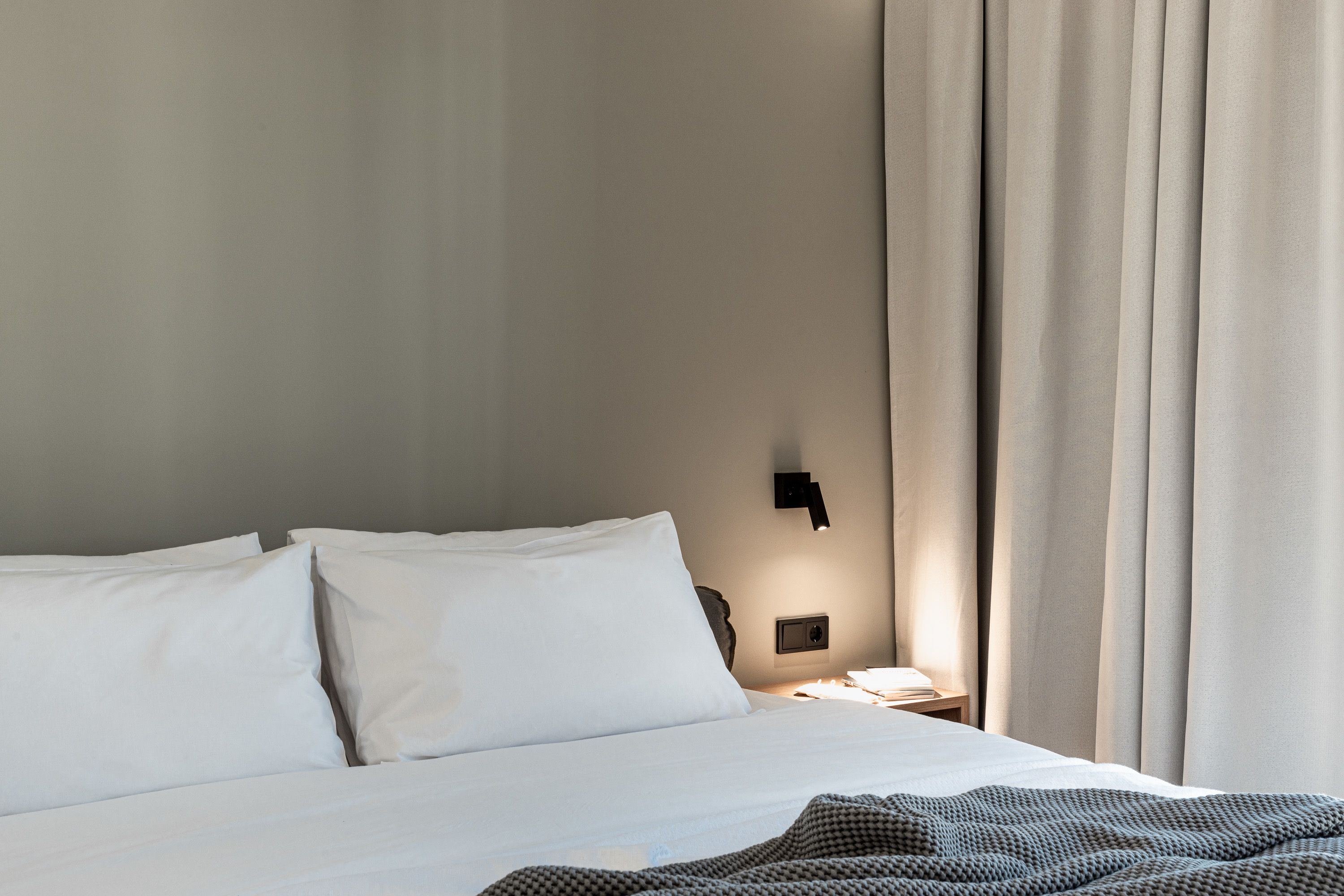 A neatly made bed with white linens, a bedside table with a lamp, and a wall-mounted light fixture.