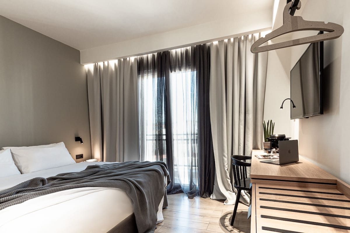 Une chambre d’hôtel moderne, équipée d’un lit, d’un bureau avec un ordinateur portable, d’un luminaire suspendu et d’une grande fenêtre avec des rideaux.