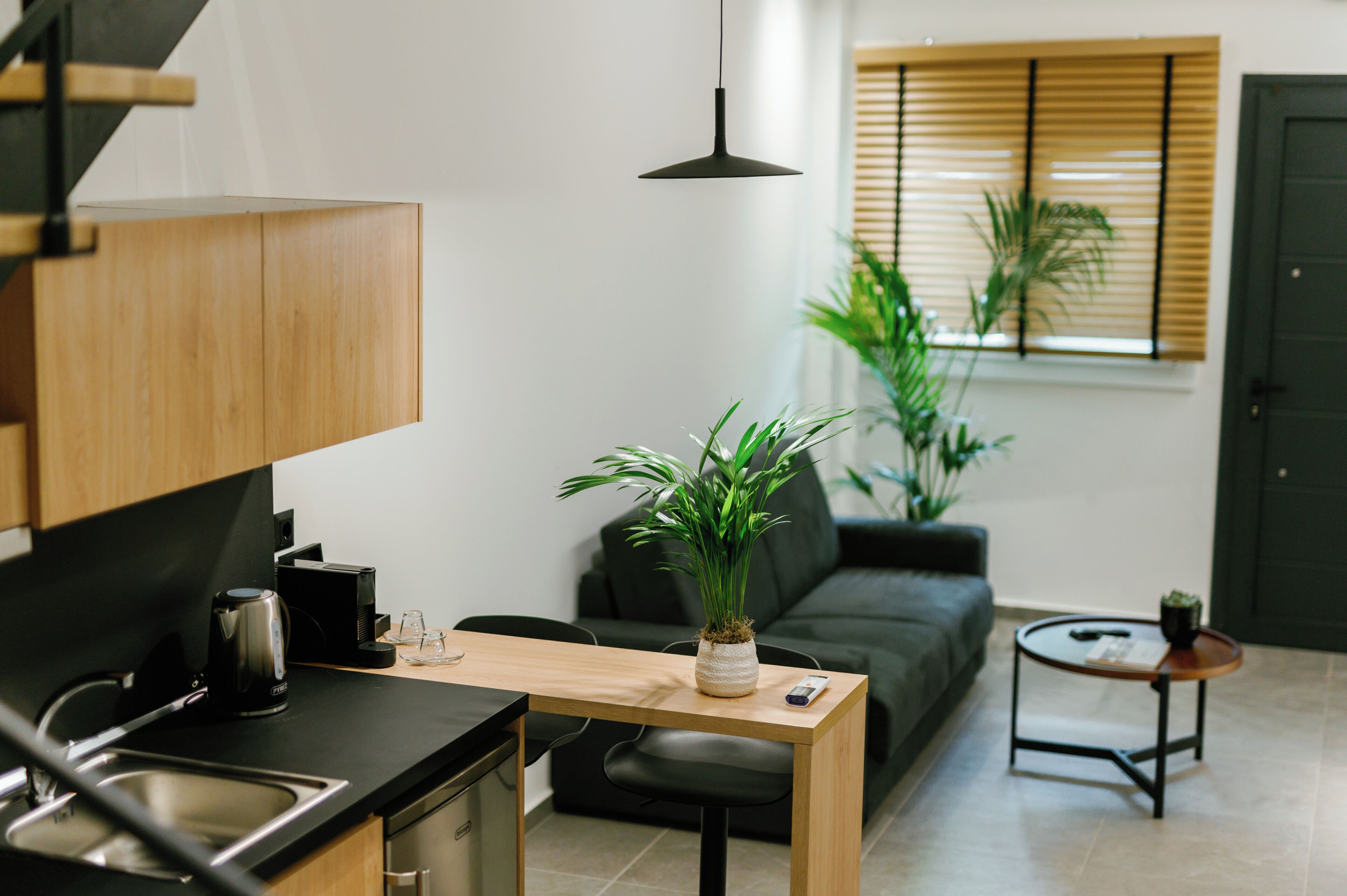 Una cocina moderna con encimera de madera, cafetera y lavabo. Hay un sofá negro y una pequeña mesa redonda con una planta.