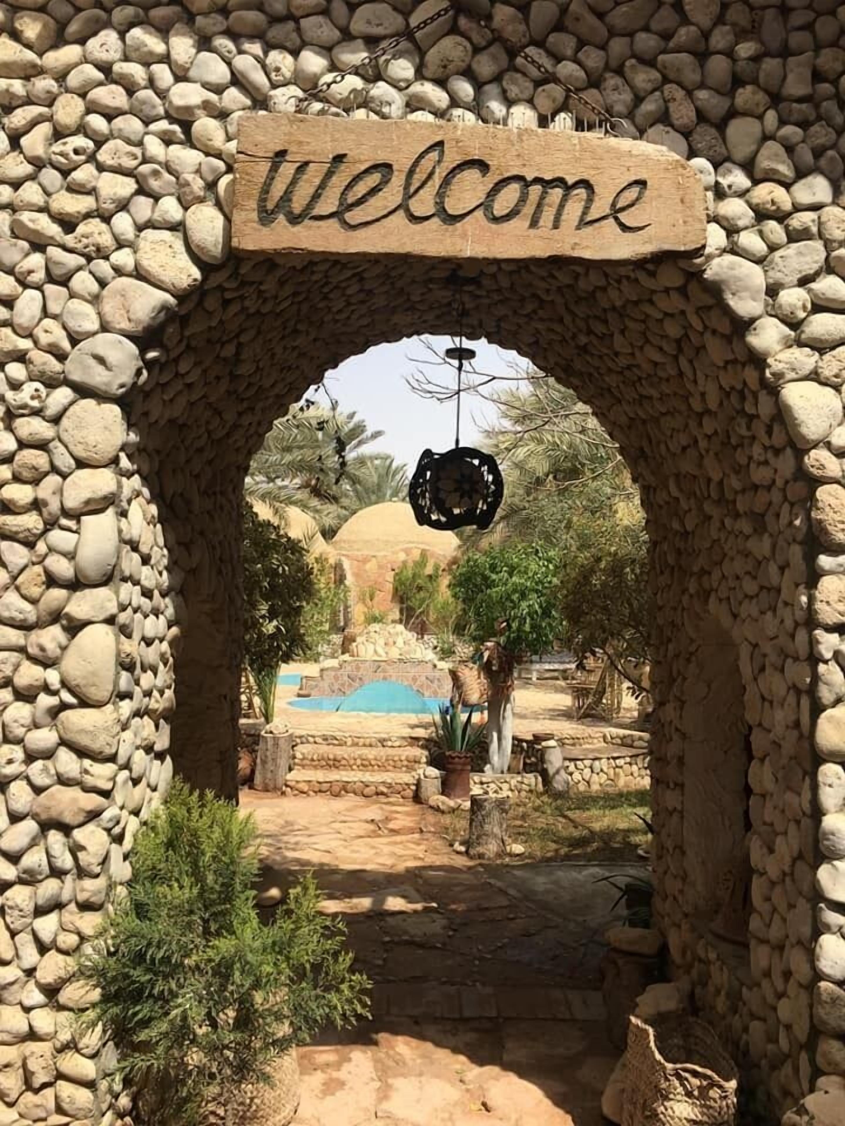 A stone archway with a 'welcome' sign, leading to a courtyard with a pool, palm trees, and a small statue.