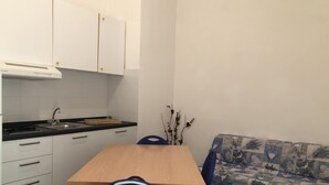 Ett litet, praktiskt kök med en spis, diskho och vita skåp. Ett litet matbord med en stol står framför köket. En mönstrad soffa står längs väggen.