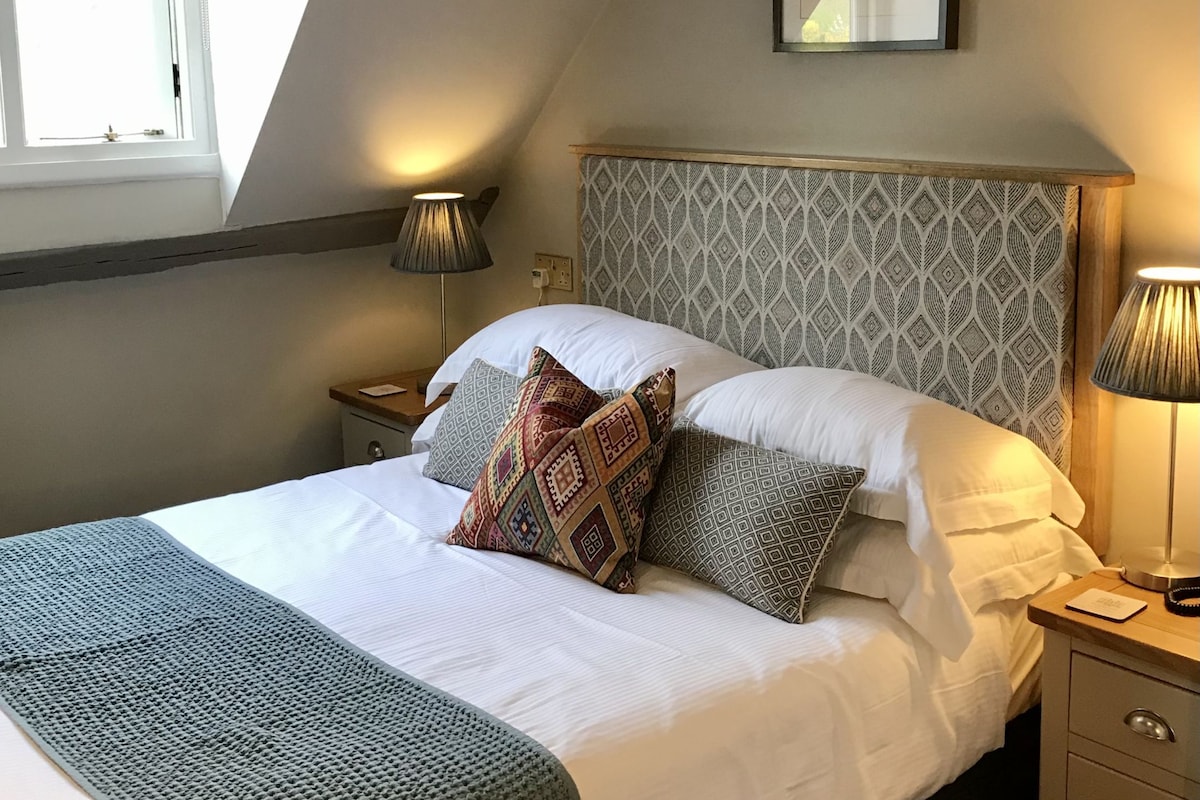 A neatly made bed with patterned pillows, a grey knitted throw, and a headboard with a geometric pattern. There are bedside lamps, a framed picture on the wall, and a window with blinds.