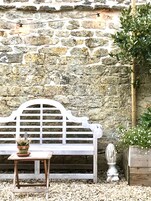 Un banco blanco con estructura metálica, una mesita con una planta en maceta y un muro de piedra con un poste de madera.