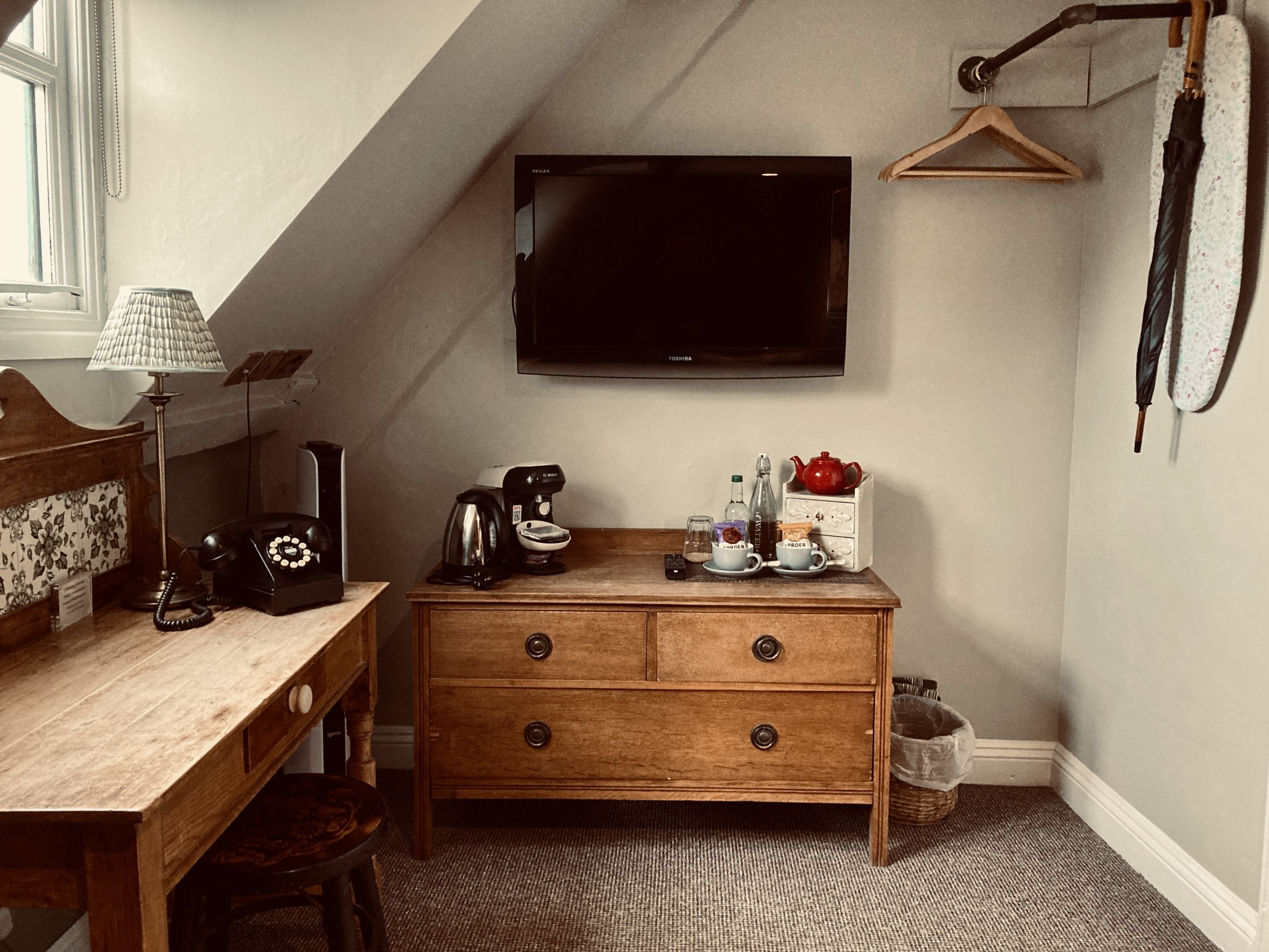 A cozy room with a wooden desk, a lamp, a coffee maker, a TV, and a hanging coat rack.
