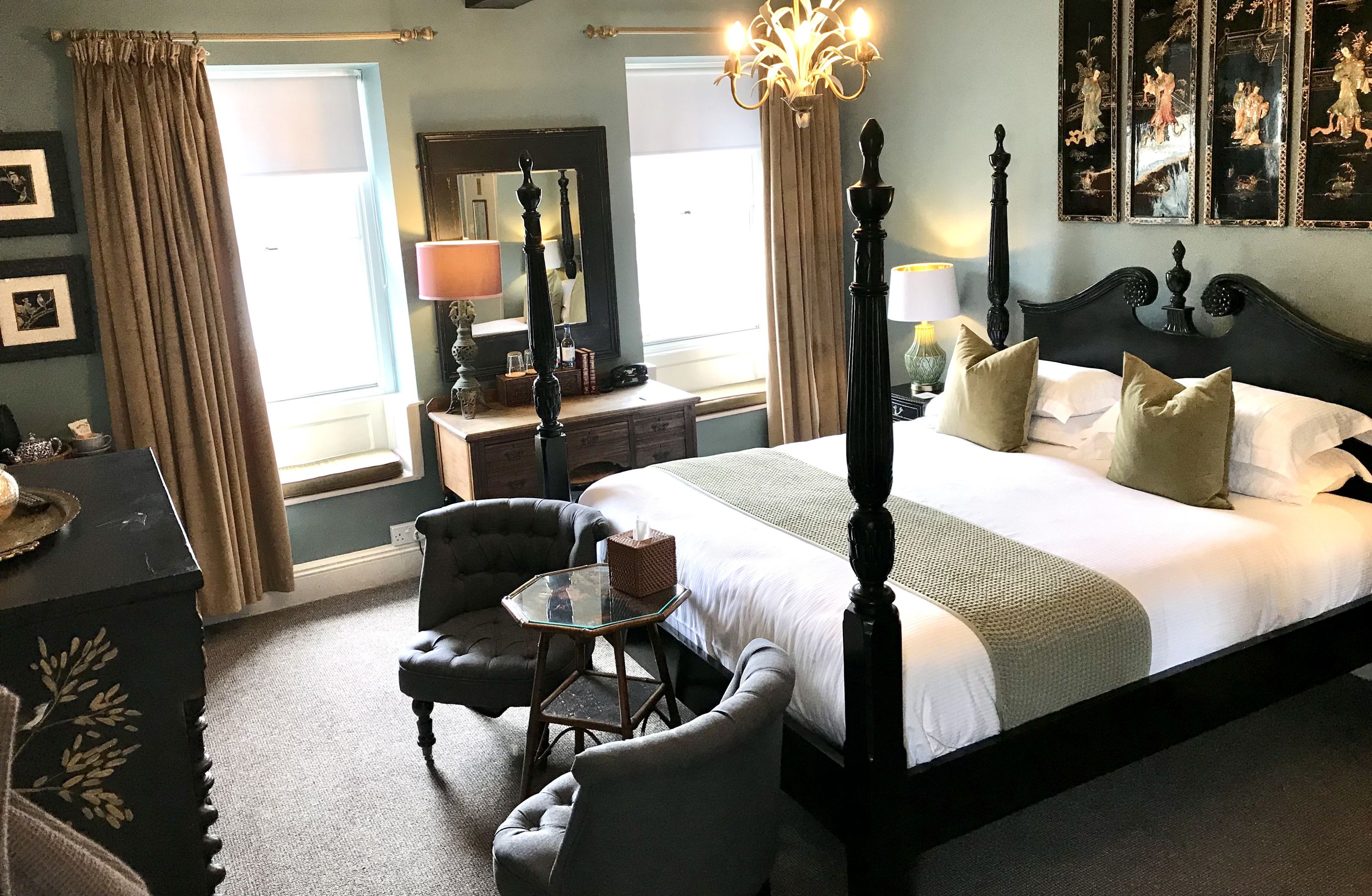 A bedroom with a four-poster bed, two armchairs, a small round table, and a dresser.