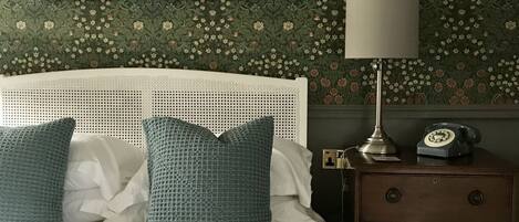 A bedroom with a white headboard, a bed with white and teal bedding, a nightstand with a lamp and a rotary phone, and floral wallpaper.