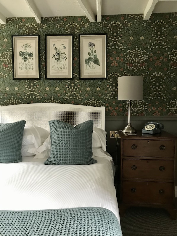 A bedroom with a white headboard, a bed with white and teal bedding, a nightstand with a lamp and a rotary phone, and floral wallpaper.