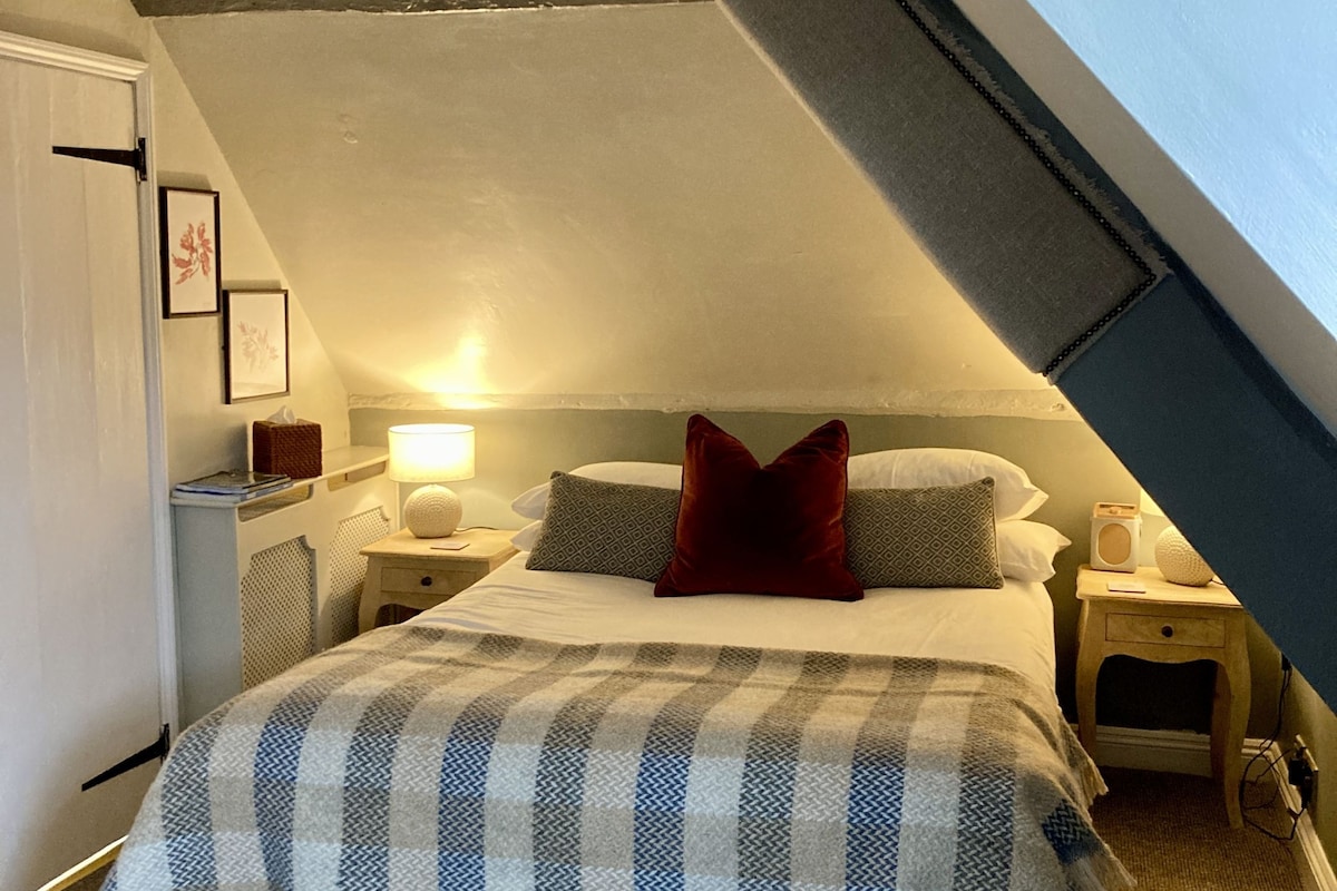 A bedroom with a sloped ceiling, a bed with a striped blanket, two bedside tables with lamps, and framed pictures on the wall.