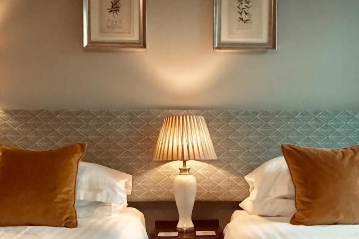 Hotellihuone, jossa on kaksi sänkyä, yöpöytä lampun kanssa ja seinällä kehystettyjä taideteoksia.