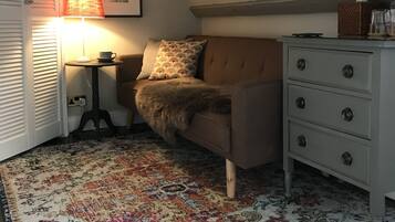 A cozy room with a patterned rug, a sofa, a small table with a lamp, a framed picture on the wall, and a dresser with drawers.