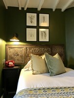 Una cama bien hecha con almohadas, un cabecero de madera con intrincados grabados, un teléfono en la mesita de noche e ilustraciones botánicas enmarcadas en la pared.