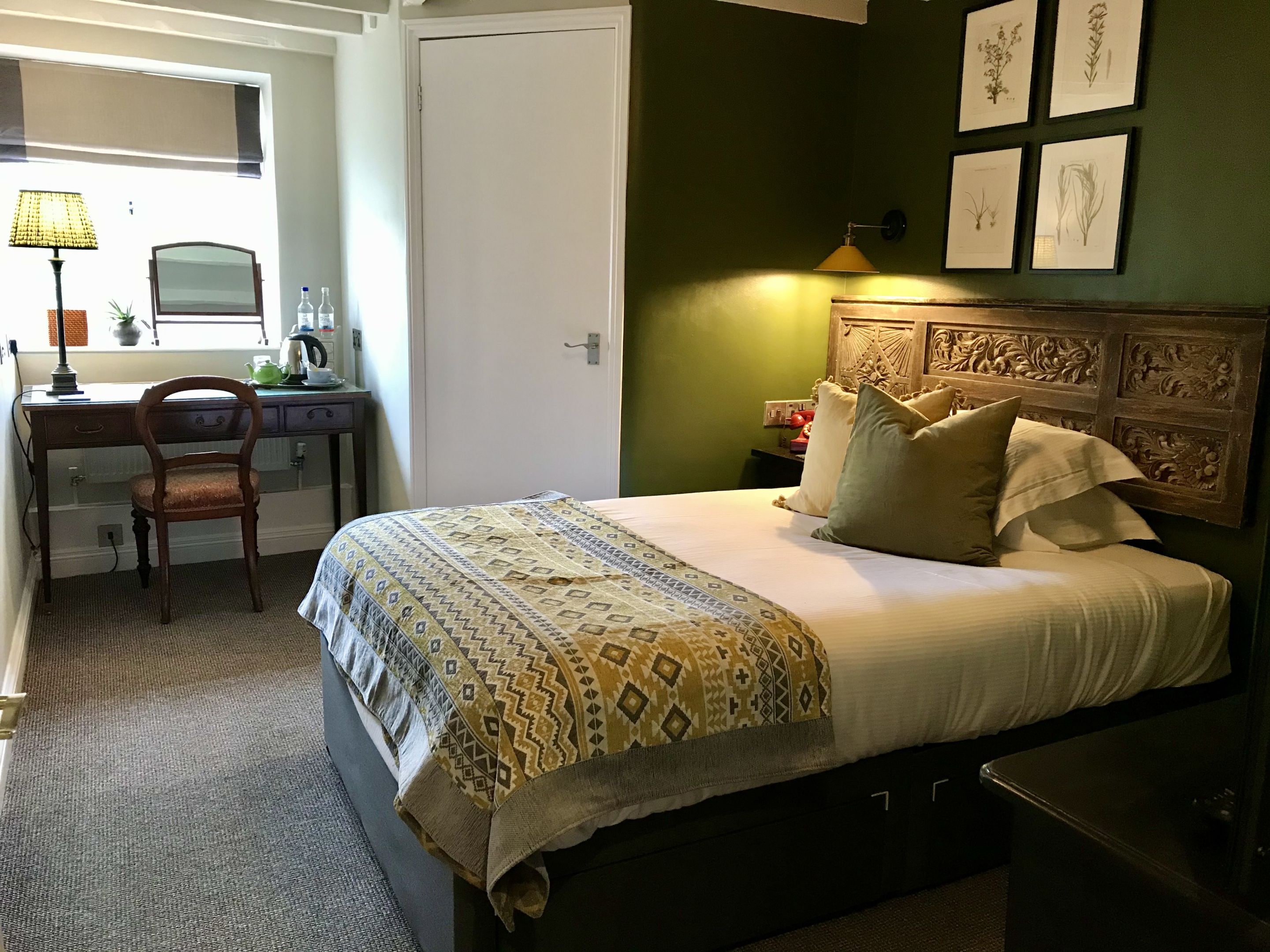 A bedroom with a bed, a desk, a chair, a lamp, and framed botanical prints on the wall.