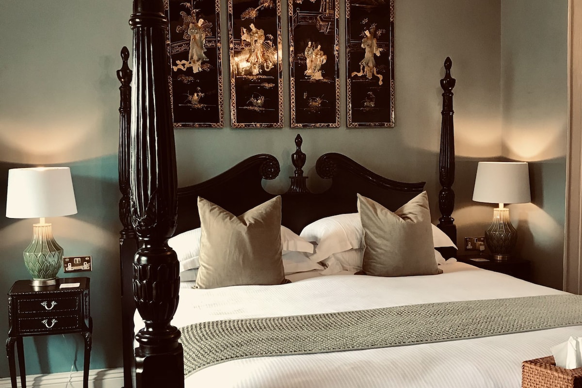 A four-poster bed with a dark finish, a set of wall-mounted paintings, two bedside tables with lamps, and a woven side table.