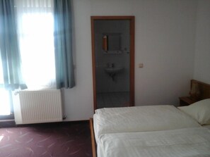 A hotel room with a bed, a radiator, a window with curtains, and a door leading to a bathroom with a sink. - Landhotel zur Krone (Krayenberggemeinde)