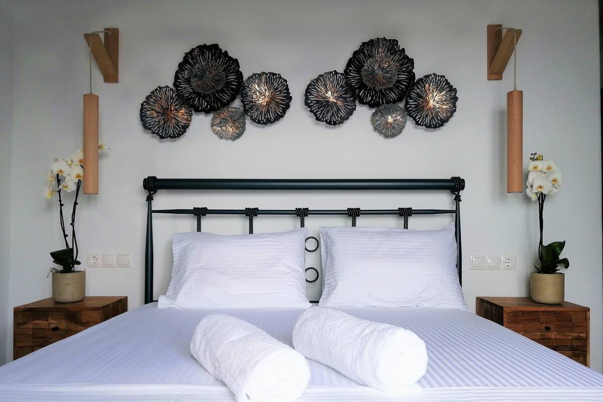 Cama bem arrumada com lençóis brancos e duas toalhas enroladas, cabeceira de metal preta e arte decorativa na parede.