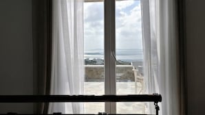 Une chambre avec un lit, une fenêtre ornée de rideaux légers et une vue sur un paysage côtier.