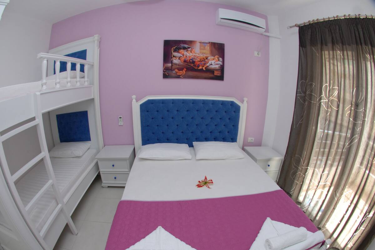A room with bunk beds, a single bed with a blue headboard, a painting on the wall, and an air conditioning unit.