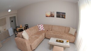 A modern living room with a L-shaped beige sofa, a matching ottoman, and a coffee table. There are two wall-mounted pictures depicting a sunset and a cityscape. - Wave Apartments Sarande (Sarandë)