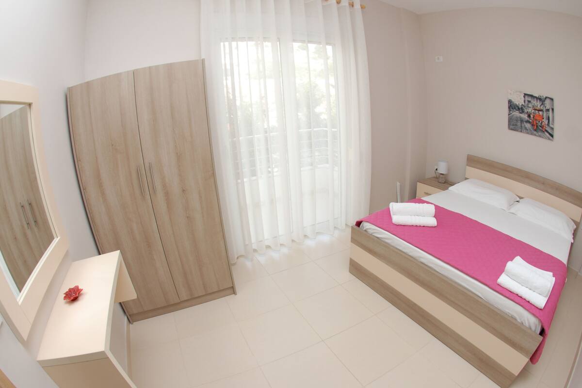 A bedroom with a bed, a bedside table, a mirror, a wardrobe, and a window with curtains.