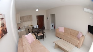 A living room with a beige sofa, a wooden coffee table, and a dining table with chairs. There is a kitchen area with cabinets and a washing machine. - Wave Apartments Sarande (Sarandë)