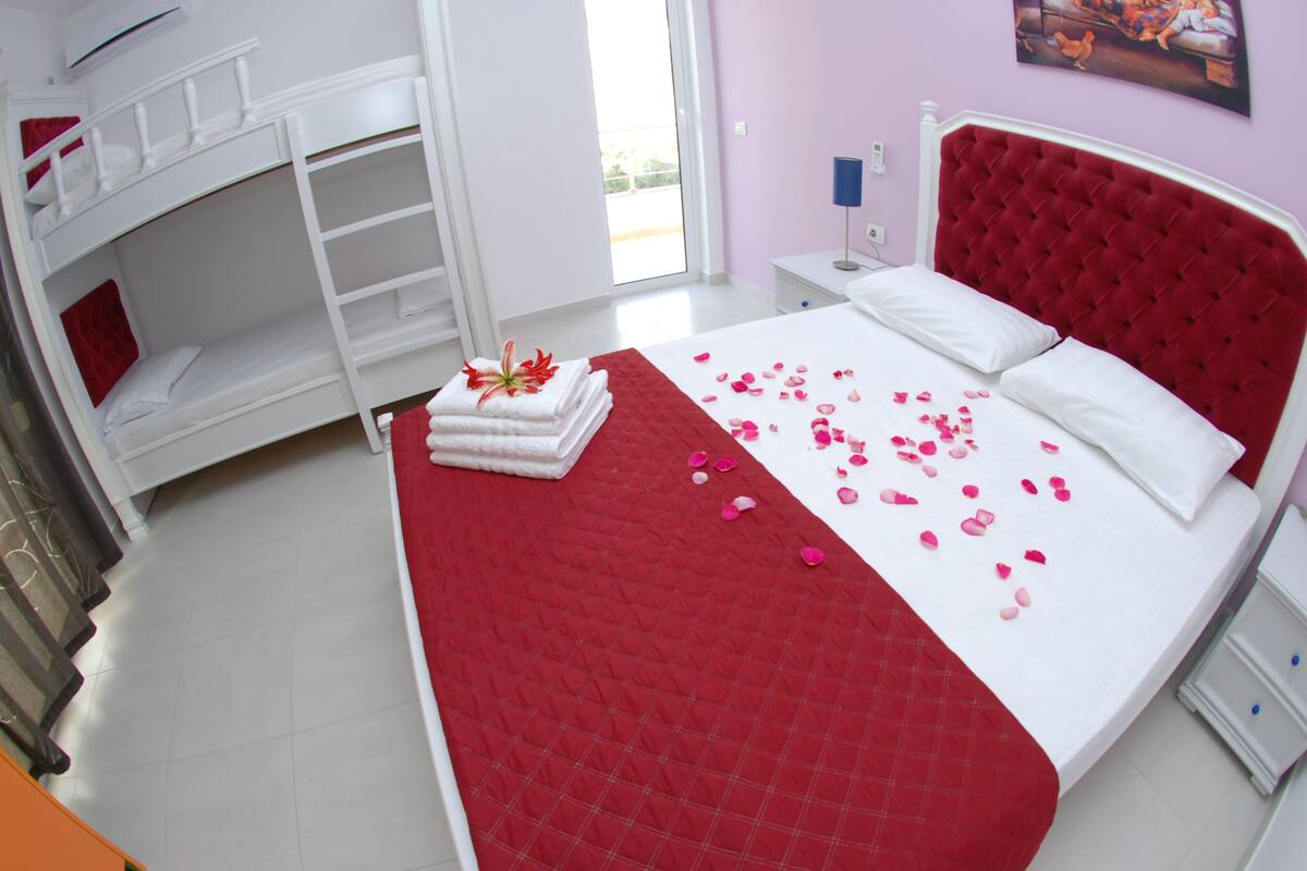 A hotel room with a large bed, a red quilted bedspread, a white headboard, and a painting on the wall.
