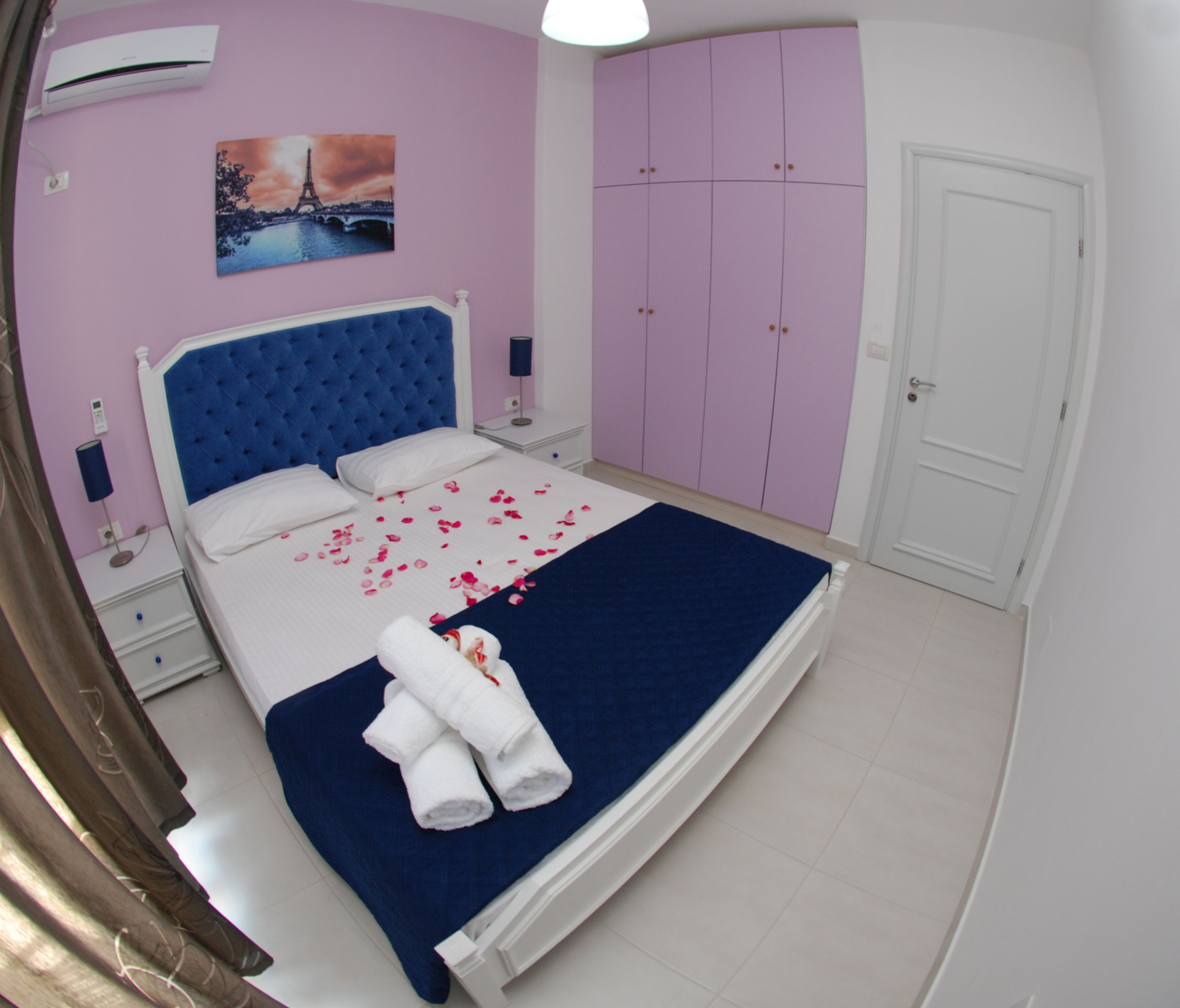 A bedroom with a large bed, a nightstand, a wall-mounted air conditioner, a door, and a pink wardrobe.