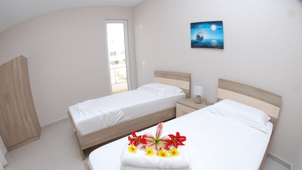 A hotel room with two single beds, a large bed with a towel and flowers, a wardrobe, a nightstand, and a painting on the wall. - Wave Apartments Sarande (Sarandë)