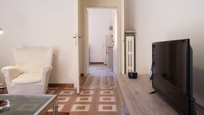 A room with a white armchair, a glass-top table with a remote control, a wooden side table with a television, and a patterned floor. - Roma - Via la Spezia 70 (Rome)