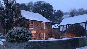 Una casa de ladrillo con tejado nevado, rodeada de árboles y un muro de piedra.