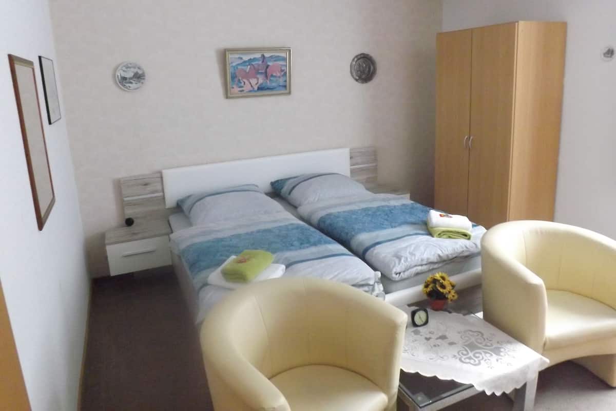 A bedroom with two beds, a wardrobe, two chairs, a small table with a clock, and a wall with two decorative plates.