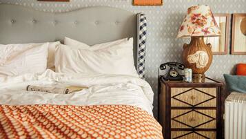 Un lit bien fait, avec du linge de lit blanc, une couverture orange à motifs, un téléphone ancien, une lampe avec un abat-jour à fleurs et une petite table sur laquelle se trouve une bouteille d’eau.