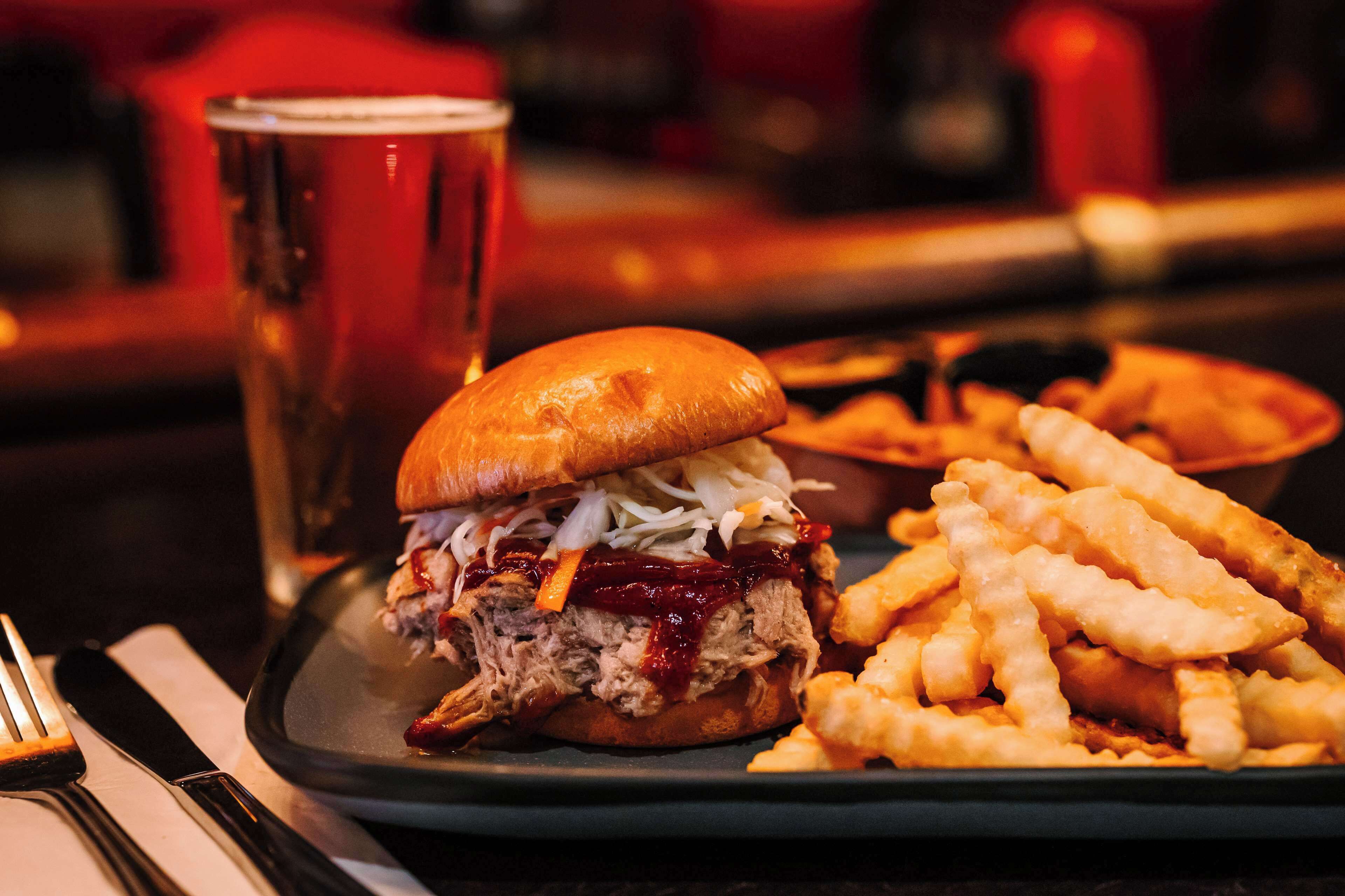 A plate with a pulled pork sandwich and fries, a glass of beer, and a fork on a table.