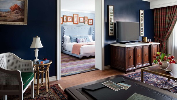 A hotel room with a bed, a television, a wooden cabinet, a wicker chair, a small table with a lamp, and a painting on the wall.