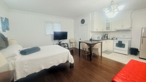 Une pièce de taille modeste comprenant un lit, un téléviseur, une table à manger avec des chaises, un coin cuisine avec une cuisinière et un four à micro-ondes, et un pouf rouge.