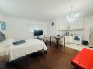 Ett litet rum med en säng, en tv, ett matbord med stolar, en köksdel med spis och mikrovågsugn samt en röd pall.
