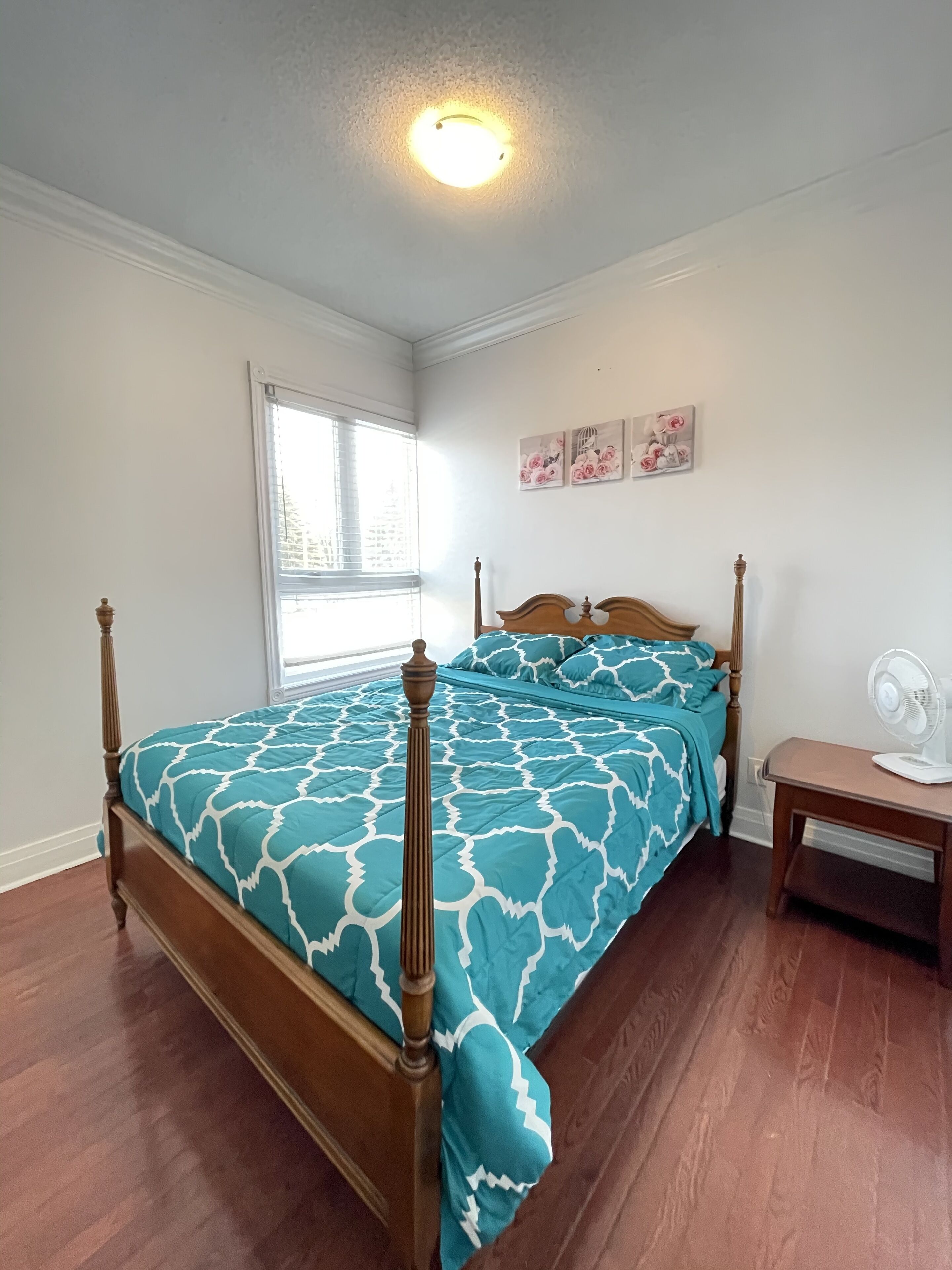 A bedroom with a wooden four-poster bed, a window with blinds, a bedside table with a fan, and floral wall art.