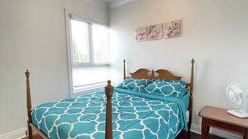 A bedroom with a wooden four-poster bed, a window with blinds, a bedside table with a fan, and floral wall art.