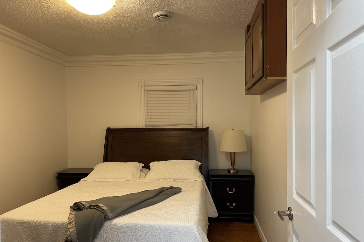 A bedroom with a bed, a nightstand with a lamp, a window with blinds, and a door.