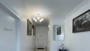 Un couloir aux murs blancs, un paillasson noir, un lustre, des tableaux encadrés et une petite table contenant du matériel de nettoyage.