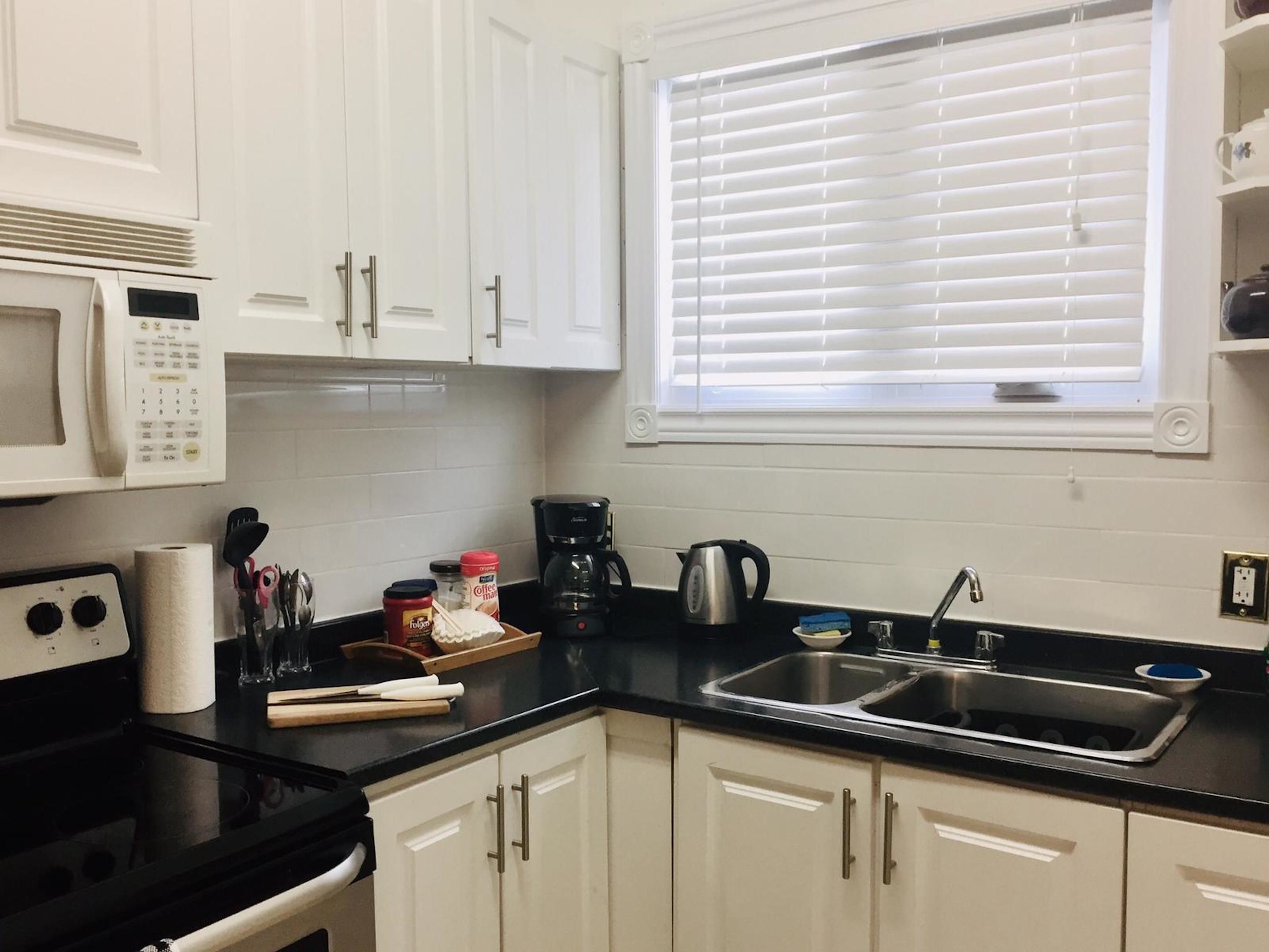 Una cocina con armarios blancos, encimera negra, cafetera, hervidor y una ventana con persianas.
