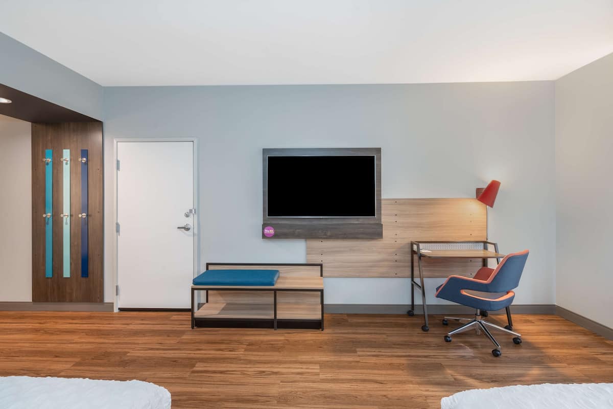 A modern room with a wooden floor, a bed, a desk with a chair, a TV mounted on the wall, and a door with a colorful panel.