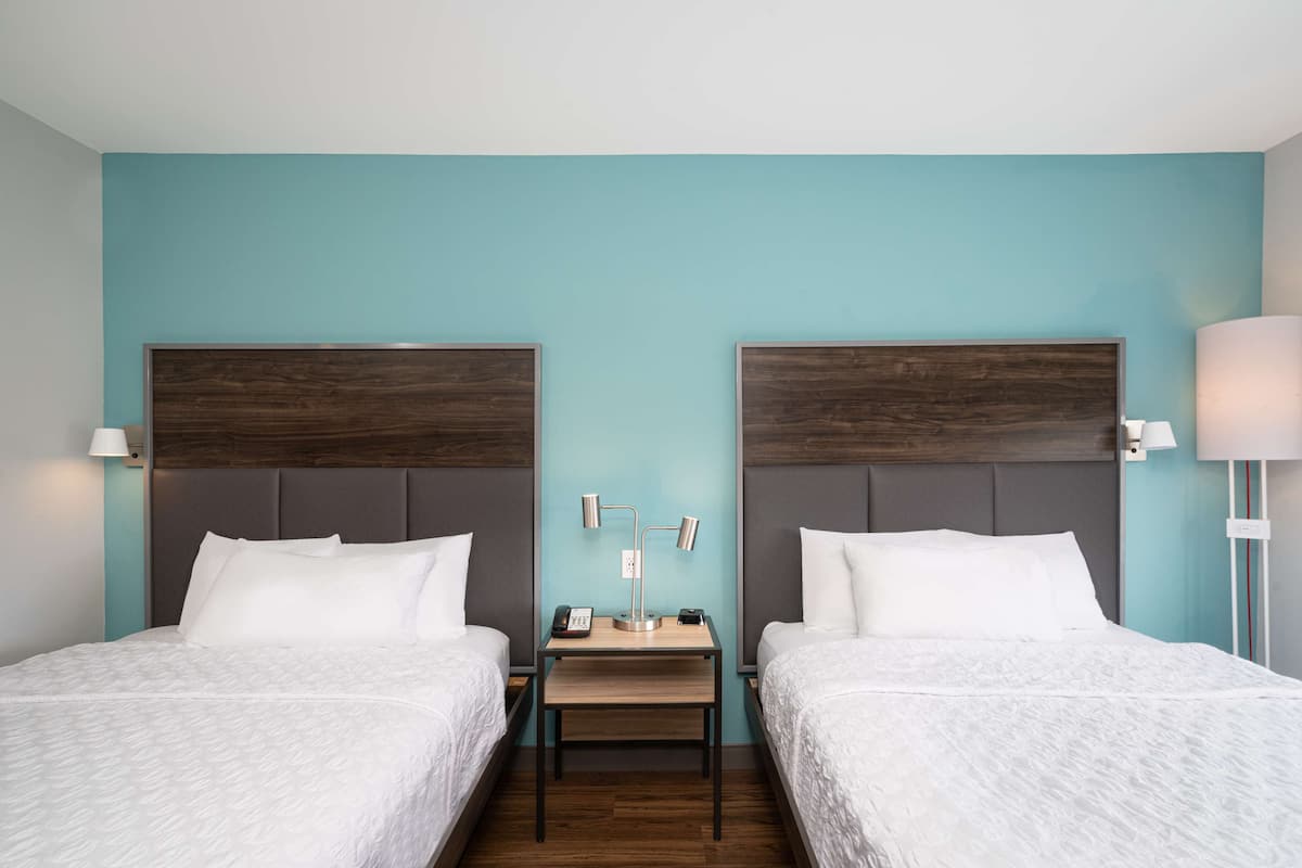 A hotel room with two beds, a small table, and a nightstand with a lamp.