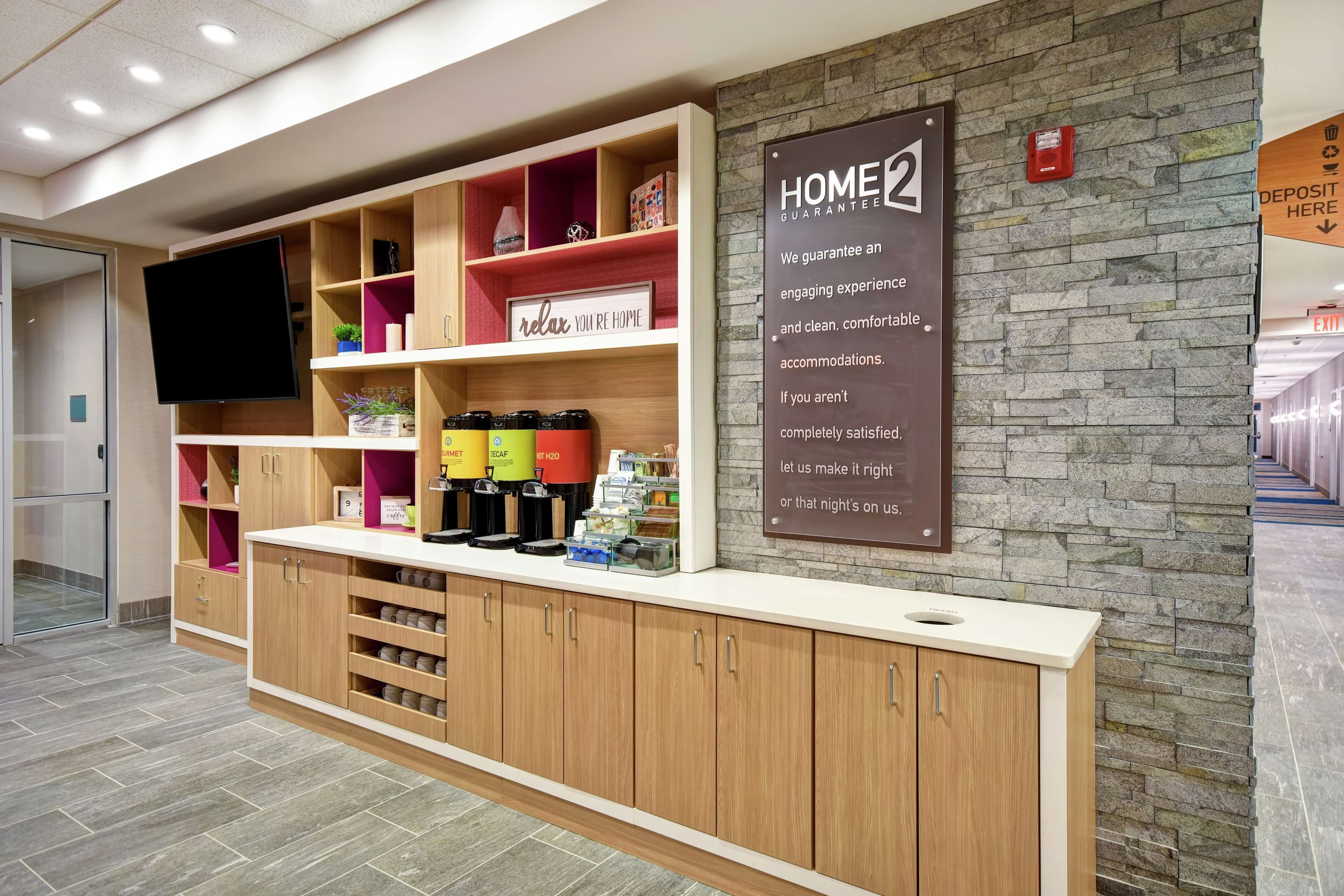 A hotel reception area with a wooden counter, shelves with amenities, a sign about home guarantee, and a stone wall.