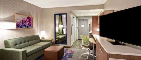 Une chambre d’hôtel avec un canapé vert, un téléviseur à écran plat, un bureau avec une chaise et un tapis coloré.