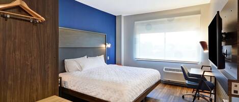 A modern hotel room with a large bed, a desk, a chair, a TV, and a window with blinds.