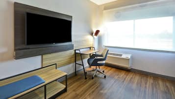 Habitación de hotel moderna con un televisor de pantalla plana grande montado en la pared, un escritorio con una silla, un banco y un ventanal con vistas al exterior.